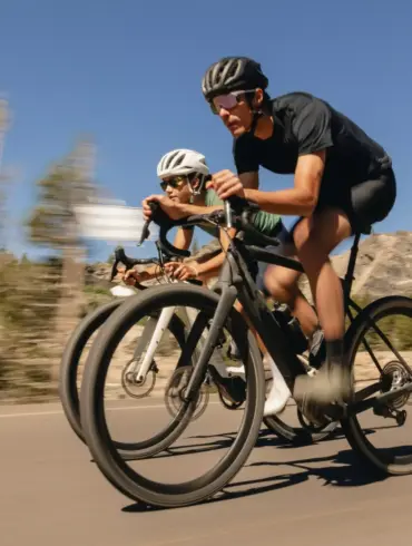 Zwei Radfahrer in sportlicher Haltung auf E-Gravelbikes im Freien mit felsigen Bergen und Bäumen im Hintergrund