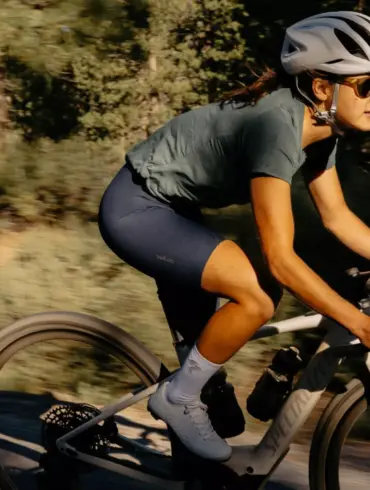 Eine Frau mit Helm und Sonnenbrille fährt ein Specialized Turbo Creo 2 Expert E-Gravelbike auf einem Schotterweg in natürlicher Umgebung.