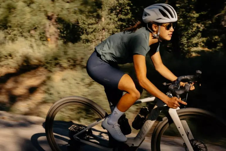 Eine Frau mit Helm und Sonnenbrille fährt ein Specialized Turbo Creo 2 Expert E-Gravelbike auf einem Schotterweg in natürlicher Umgebung.