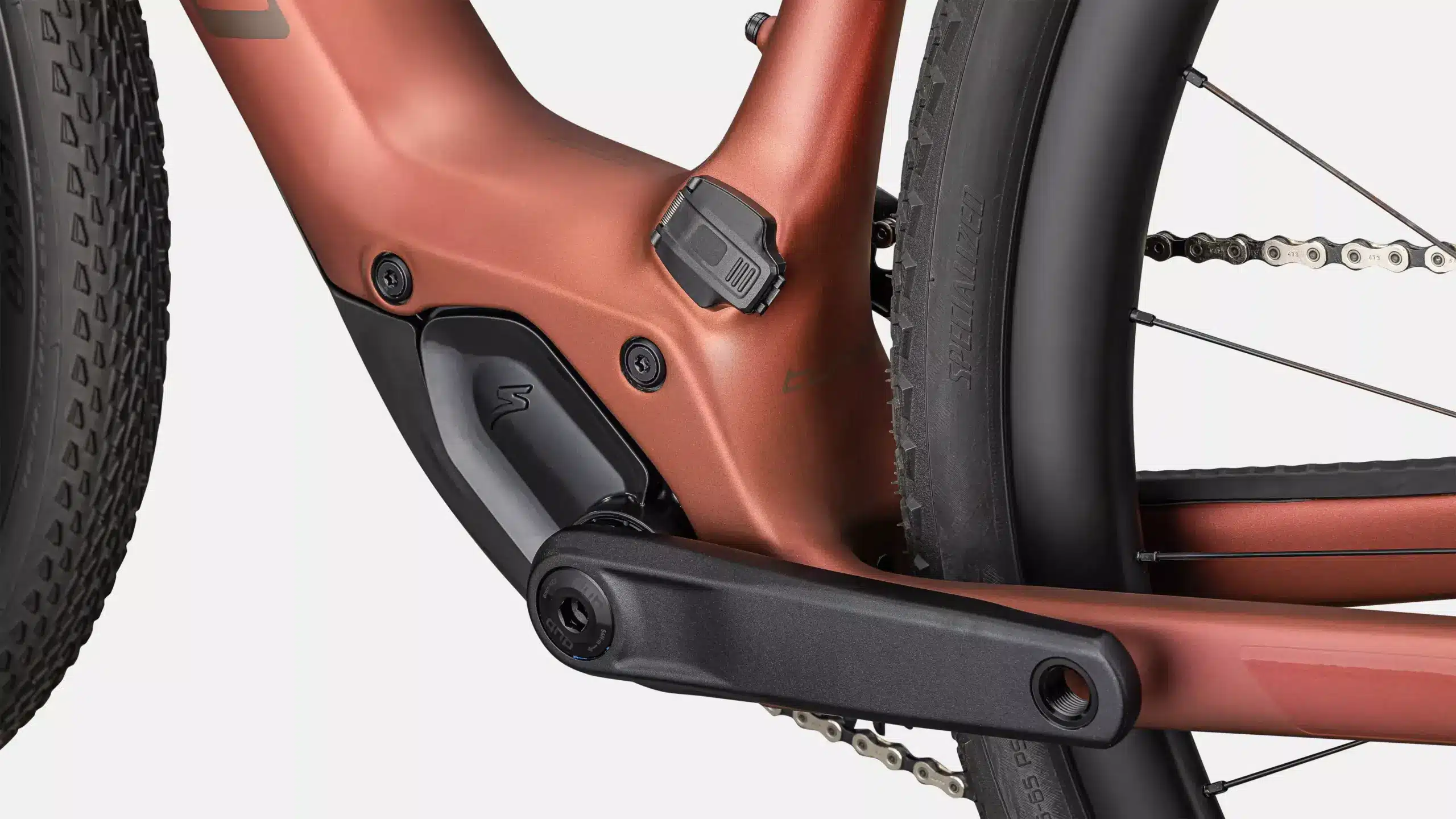 Nahaufnahme des Tretlagerbereichs eines orangefarbenen E-Gravelbikes mit schwarzer Kurbel, Kette und Teilen des Hinterrads mit Reifen.