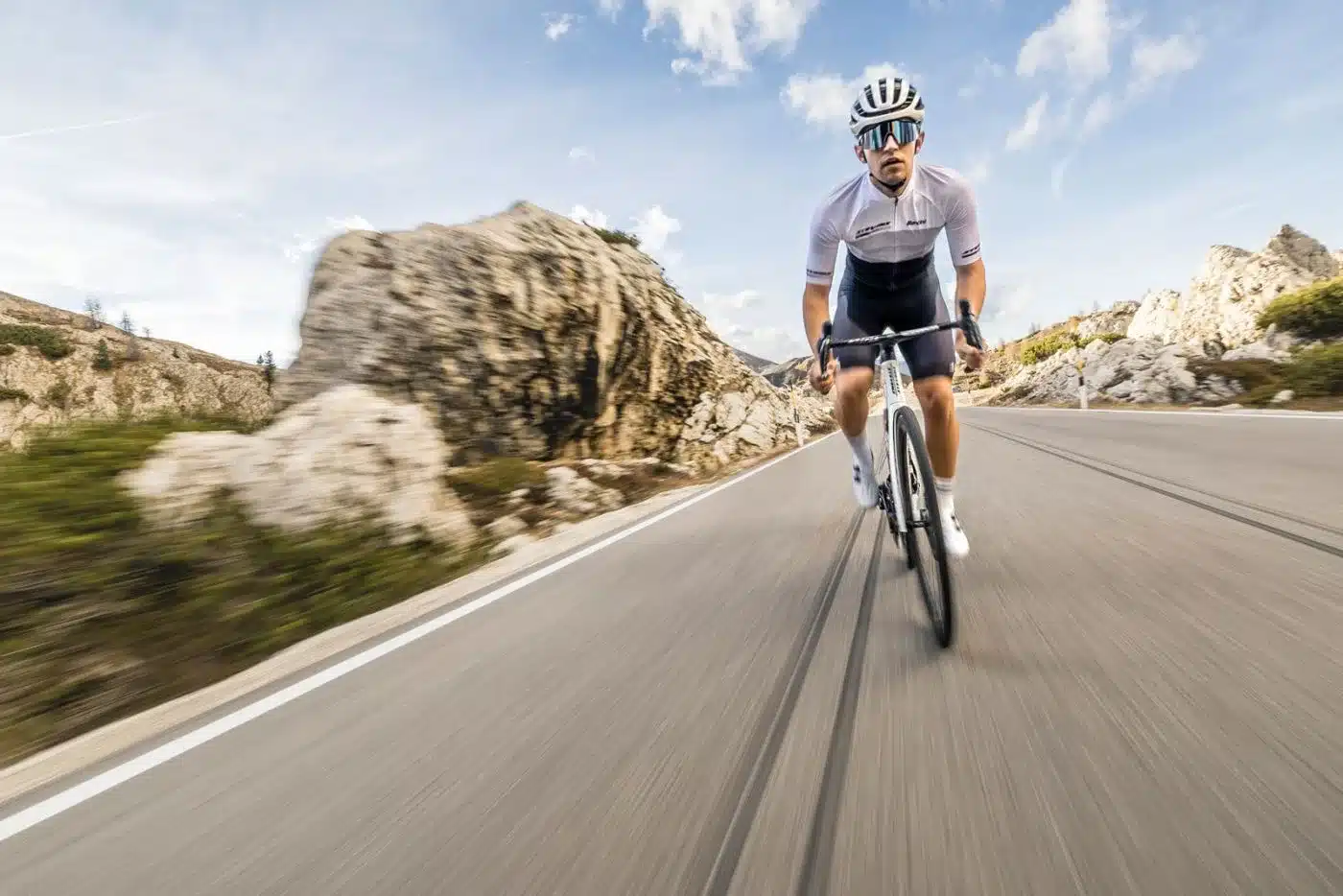 Ein Mann in voller Radsportmontur fährt auf einem weißen Stevens Arcalis Rennrad auf einer asphaltierten Straße in den Bergen bergab, mit Felsen und Büschen auf beiden Seiten und blauem Himmel mit Wolken darüber.