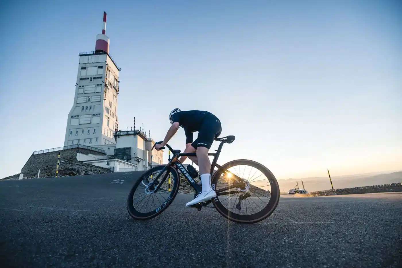 Radsportler in voller Montur fährt ein Stevens Arcalis Aero-Rennrad auf einer asphaltierten Straße bergauf, mit einer großen, weißen Sendeanlage im Hintergrund und Sonnenschein.