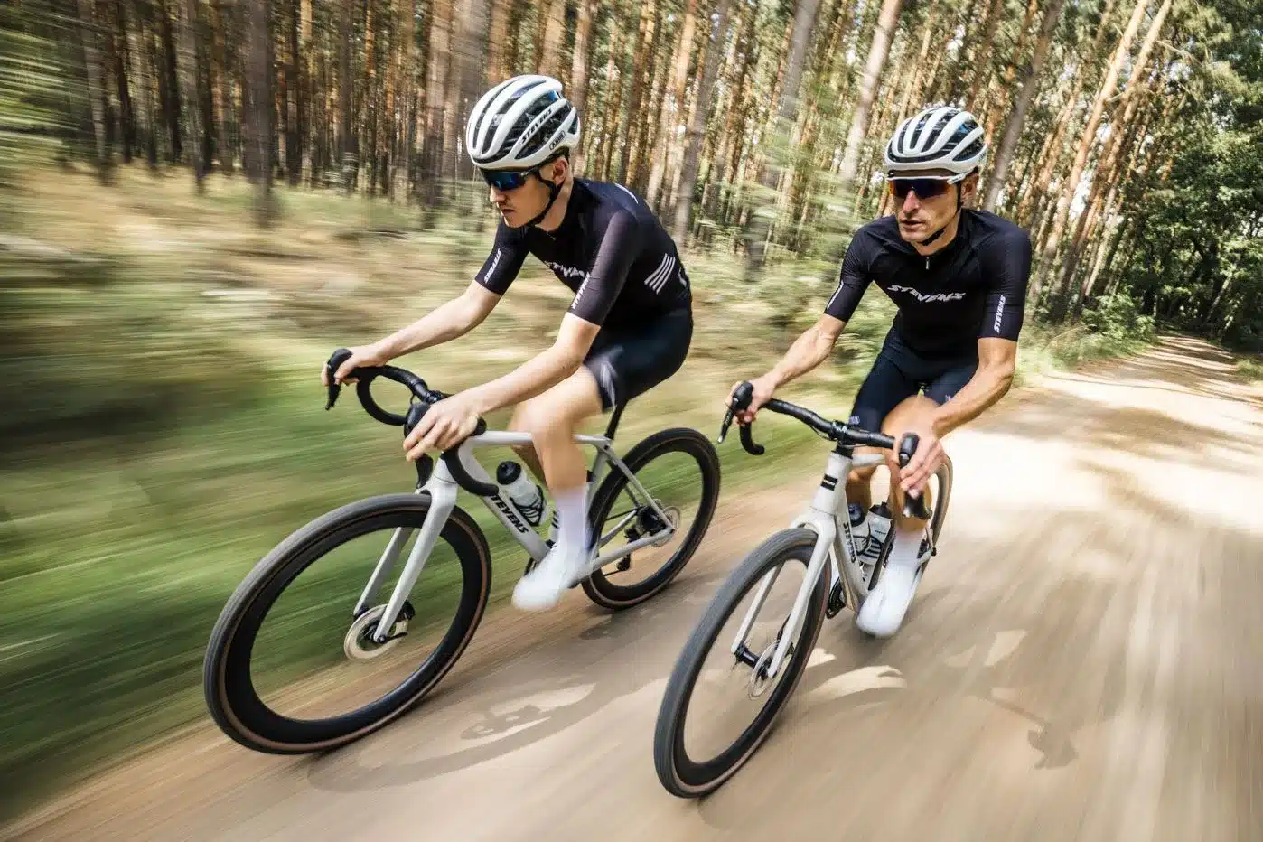 Zwei Männer fahren nebeneinander auf einem Gravel-Racebike durch einen Waldweg. Sie tragen Radsportbekleidung und Helme.