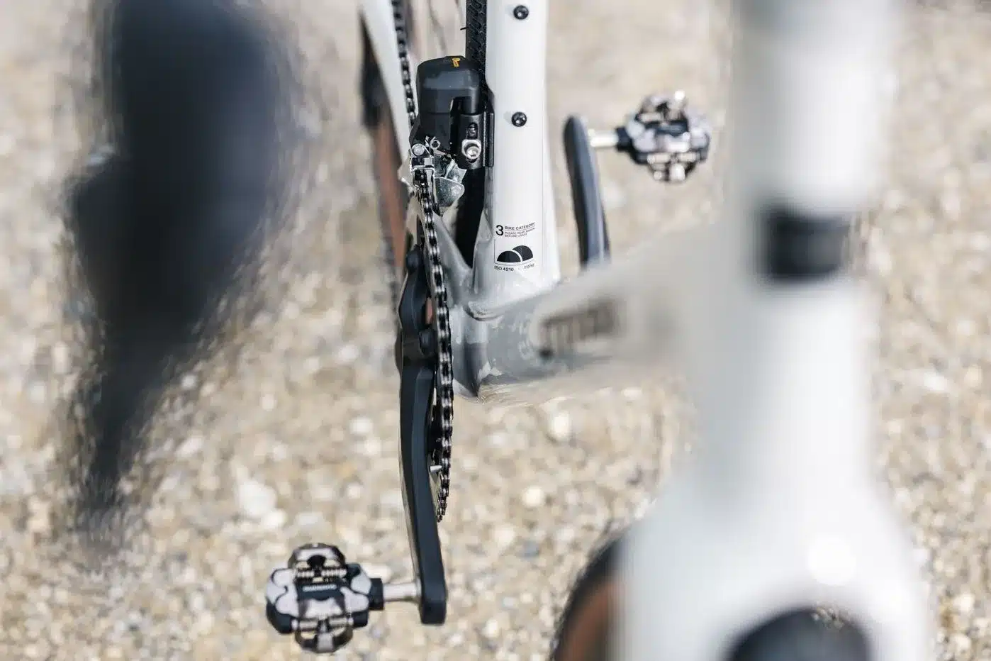 Detailaufnahme des Hinterbaus eines weißen Gravelbikes mit Carbonrahmen, zeigt die Kette, ein Pedal und die Fahrradkategorie-Aufkleber am Rahmen.