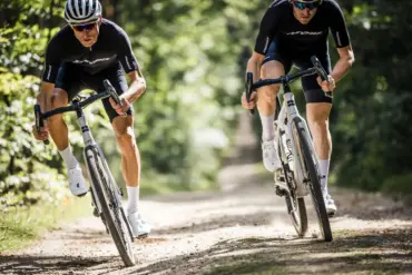 Zwei Fahrer auf modernen Gravelbikes, die auf einem Schotterweg in leicht gebückter Rennposition fahren.
