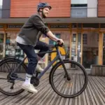 Ein Mann mit Helm fährt ein dunkles Stevens Kalea 6.1 LT Fahrrad auf einer Holzplankenoberfläche vor einem Gebäude mit Fenstern.