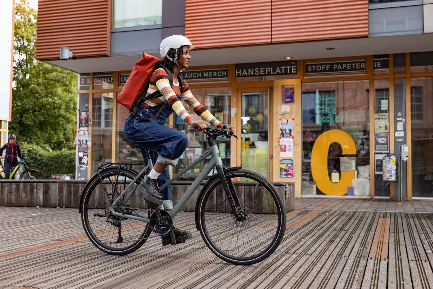 Frau mit Helm und Rucksack fährt ein graues Stevens Kalea 7.1 LT Urban Bike auf einem Holzweg vor einem Geschäft.