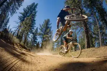 Zwei Personen fahren mit E-Gravelbikes auf einem staubigen Waldweg. Eine Person macht einen Sprung.