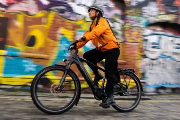 Eine Frau mit Helm und orangefarbener Jacke fährt ein schwarzes Specialized Turbo Vado 4.0 E-Bike auf einer Straße mit Graffitimauer im Hintergrund.