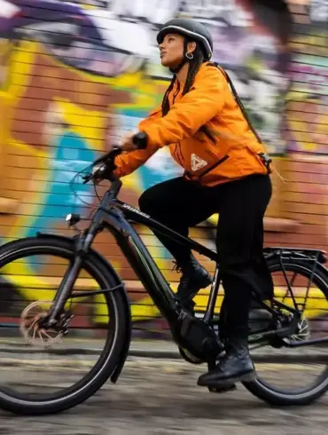 Eine Frau mit Helm und orangefarbener Jacke fährt ein schwarzes Specialized Turbo Vado 4.0 E-Bike auf einer Straße mit Graffitimauer im Hintergrund.