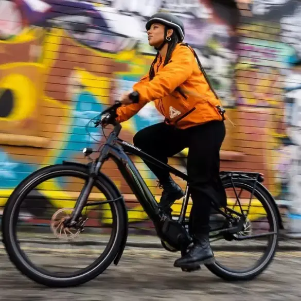 Eine Frau mit Helm und orangefarbener Jacke fährt ein schwarzes Specialized Turbo Vado 4.0 E-Bike auf einer Straße mit Graffitimauer im Hintergrund.