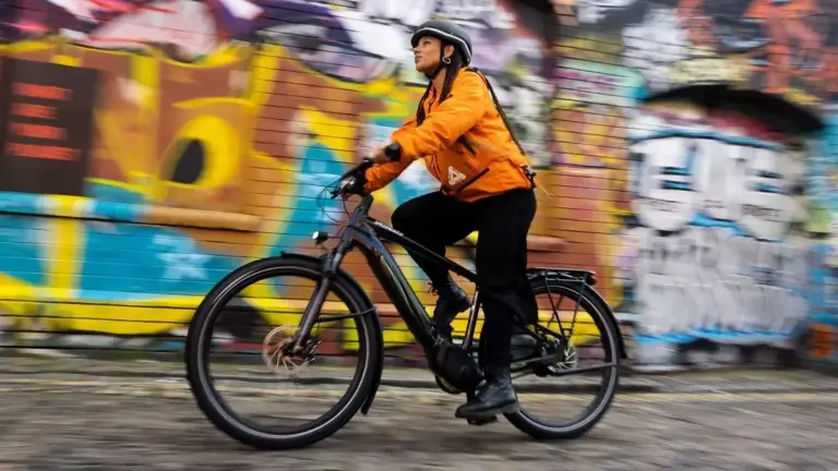 Eine Frau mit Helm und orangefarbener Jacke fährt ein schwarzes Specialized Turbo Vado 4.0 E-Bike auf einer Straße mit Graffitimauer im Hintergrund.