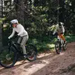 Zwei Personen fahren mit ihren E-Bikes auf einem erdigen Weg durch einen Wald mit vielen Bäumen und Grünpflanzen.