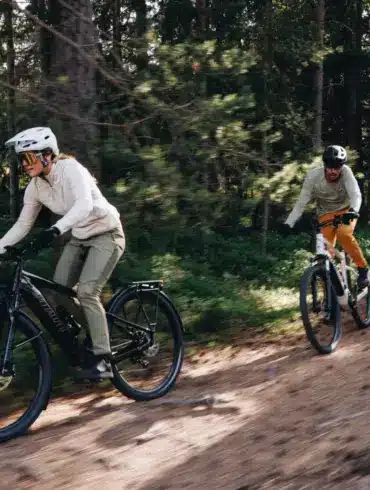 Zwei Personen fahren mit ihren E-Bikes auf einem erdigen Weg durch einen Wald mit vielen Bäumen und Grünpflanzen.
