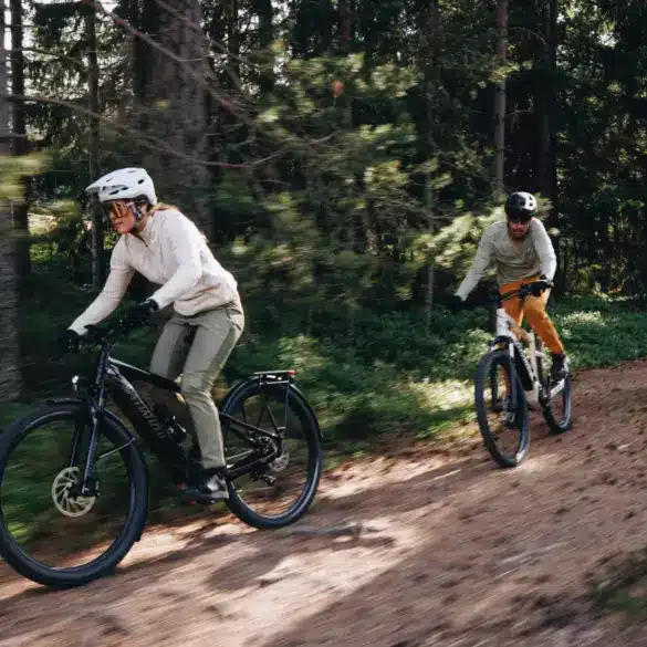 Zwei Personen fahren mit ihren E-Bikes auf einem erdigen Weg durch einen Wald mit vielen Bäumen und Grünpflanzen.