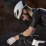 Ein Mann in aerodynamischer Rennradposition trägt einen weißen Giant Pursuit Aero Helm, schwarzes Kurzarmtrikot und weiße Handschuhe. Der Fokus liegt auf dem Helm und dem Gesicht des Fahrers, während der Lenker und die oberste Gabel des Fahrrads im unteren Bereich zu sehen sind.