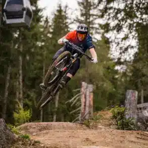 Mountainbiker in Aktion beim Sprung auf einer Mountainbike-Strecke im Wald, mit einer Seilbahn im Hintergrund.