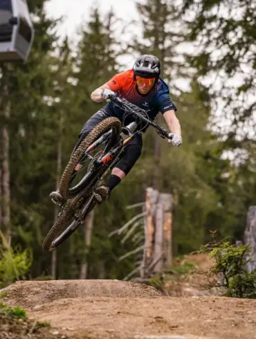 Mountainbiker in Aktion beim Sprung auf einer Mountainbike-Strecke im Wald, mit einer Seilbahn im Hintergrund.
