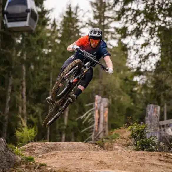 Mountainbiker in Aktion beim Sprung auf einer Mountainbike-Strecke im Wald, mit einer Seilbahn im Hintergrund.