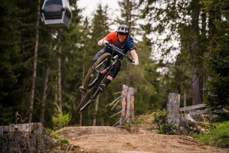 Mountainbiker in Aktion beim Sprung auf einer Mountainbike-Strecke im Wald, mit einer Seilbahn im Hintergrund.