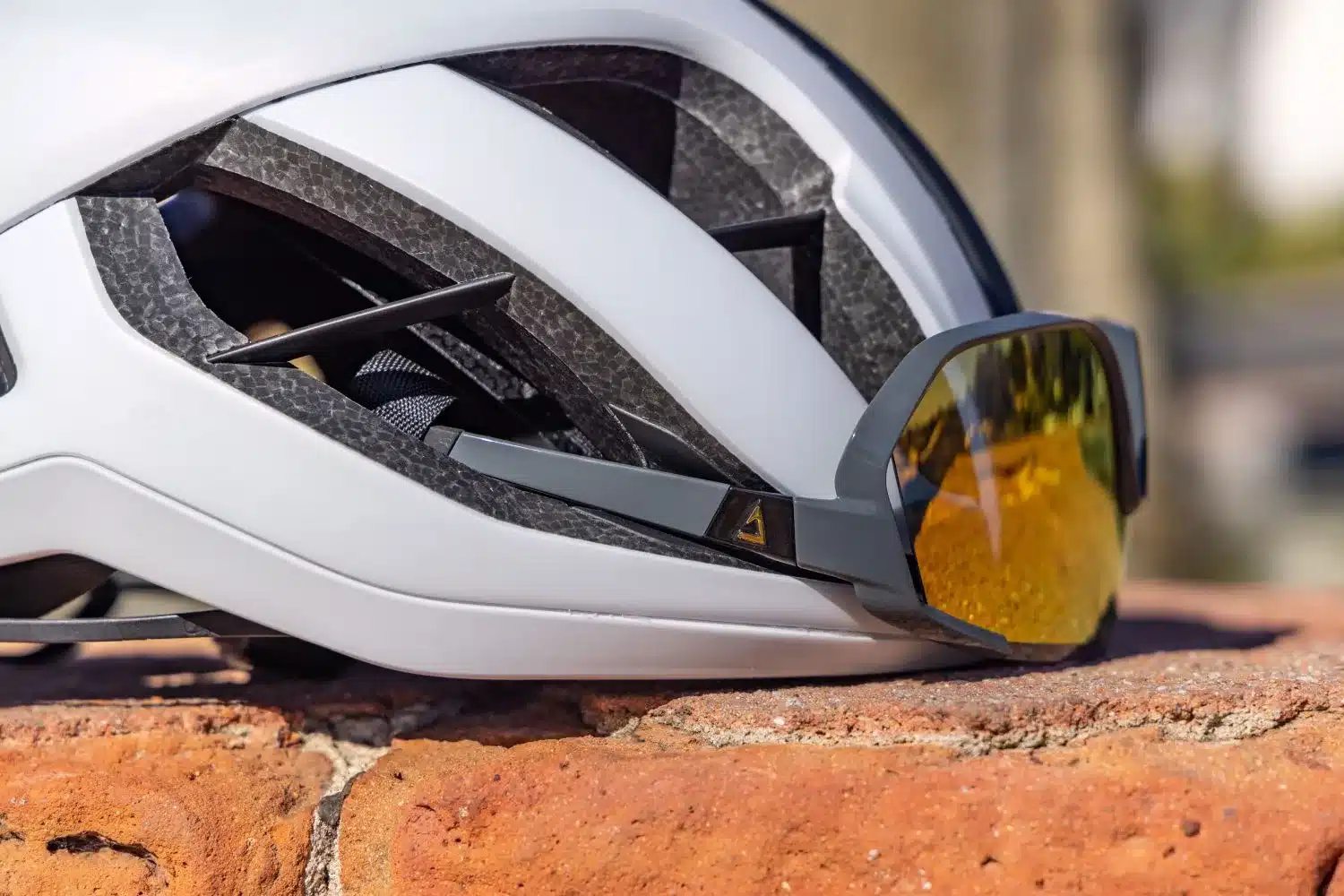 Nahaufnahme eines weißen Giant Rev Pro Rennradhelms mit einer grauen Sportbrille mit orange verspiegelten Gläsern, die in die vorderen Belüftungsöffnungen eingehakt ist.