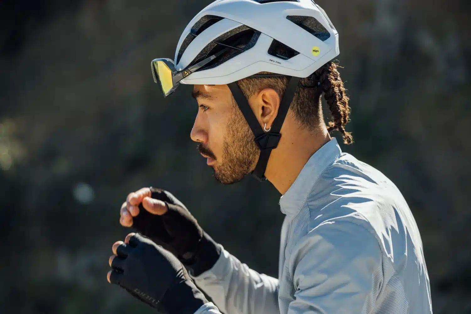 Ein Mann mit geflochtenen Haaren und einem Kinnbart trägt einen weißen Giant Rev Pro Fahrradhelm mit Mips-Technologie, der mit einer speziellen Brille ausgestattet ist.