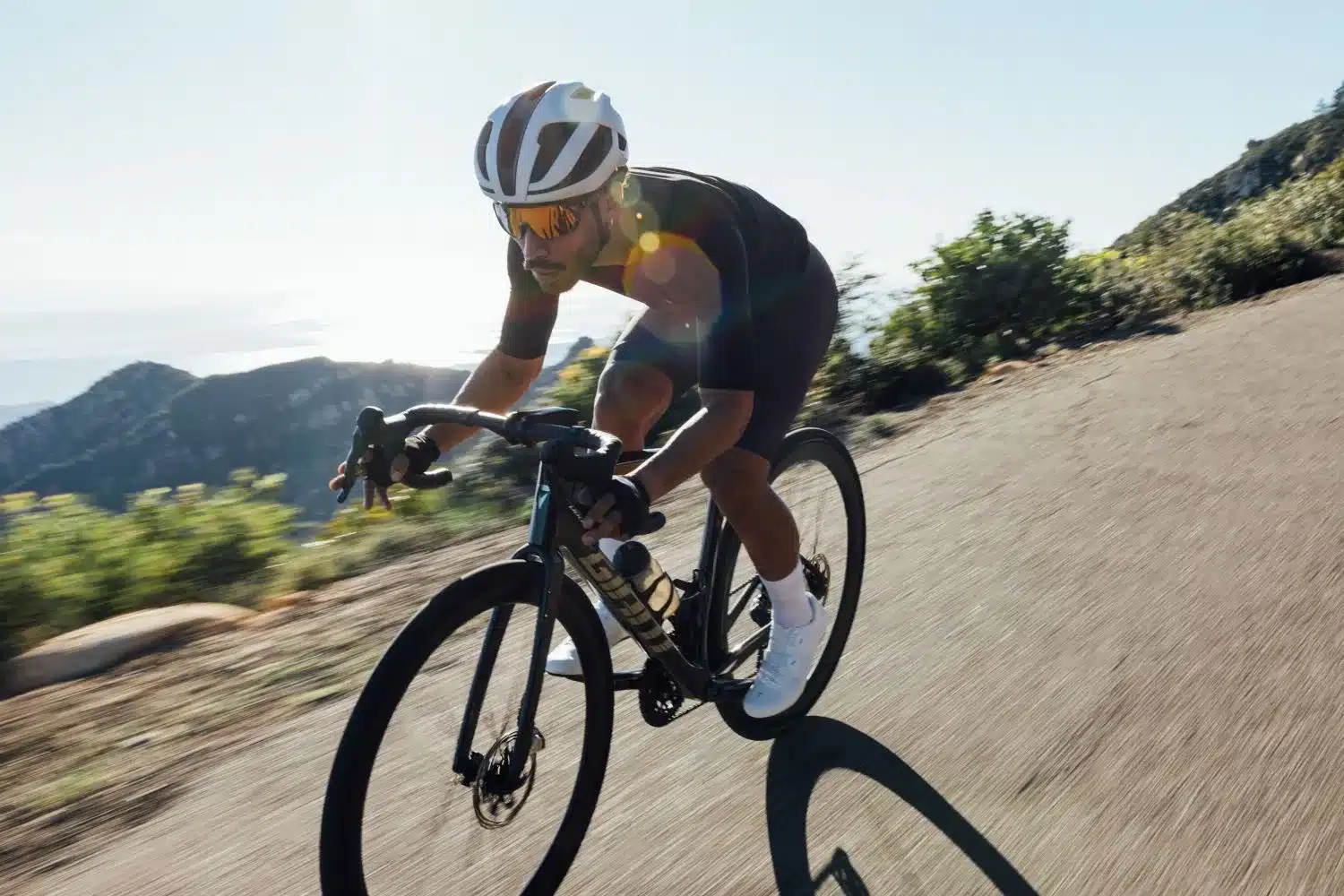 Ein Mann fährt mit einem Rennrad auf einer Bergstraße, trägt einen weißen und braunen Fahrradhelm, sportliche Kleidung und eine Sonnenbrille.