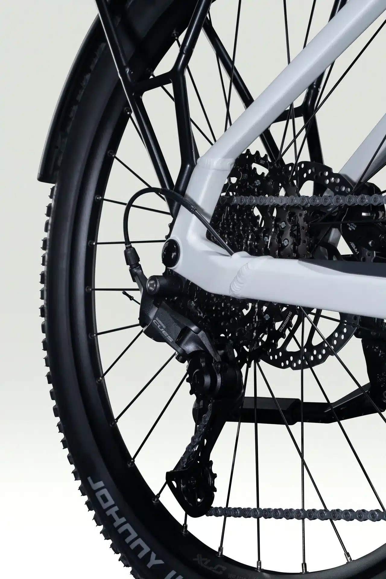 Nahaufnahme des Hinterrads eines weißen E-Trekkingbikes mit schwarzem Gepäckträger und Schutzblech. Deutlich sichtbar ist die Shimano CUES 1x11 Schaltung mit einer 11-50 Kassette und die breite Bereifung 'Johnny Watts'.