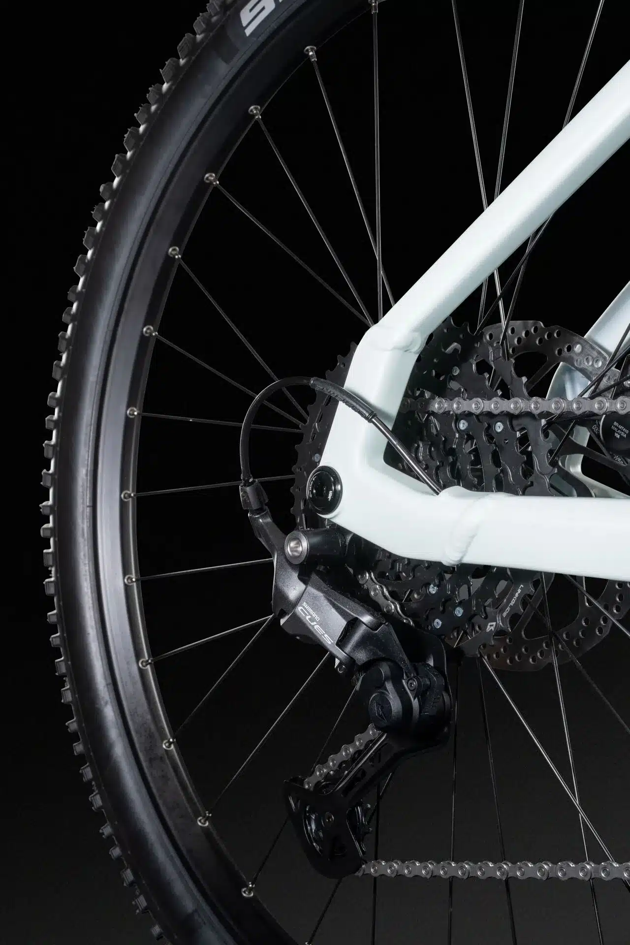 Nahaufnahme des Hinterrads eines weißen E-Mountainbikes, die das schwarze Schaltwerk, die Kette und die Kassette zeigt. Der Reifen mit Stollenprofil ist ebenfalls sichtbar.