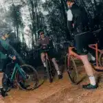 Drei Radfahrer stehen mit ihren Fahrrädern auf einem erdigen Weg im Wald, umgeben von Bäumen. Einer der Radfahrer im Vordergrund hat ein orangefarbenes Fahrrad. Im Hintergrund stehen zwei weitere Radfahrer mit ihren Fahrrädern.