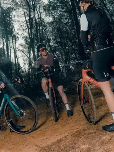 Drei Radfahrer stehen mit ihren Fahrrädern auf einem erdigen Weg im Wald, umgeben von Bäumen. Einer der Radfahrer im Vordergrund hat ein orangefarbenes Fahrrad. Im Hintergrund stehen zwei weitere Radfahrer mit ihren Fahrrädern.