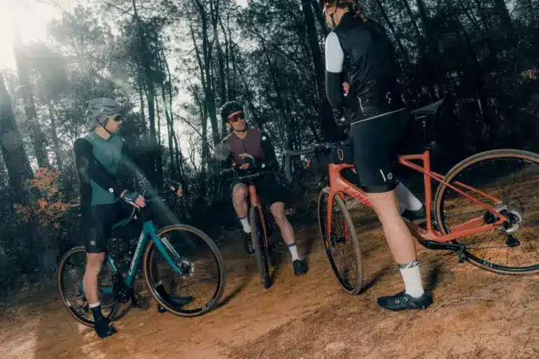 Drei Radfahrer stehen mit ihren Fahrrädern auf einem erdigen Weg im Wald, umgeben von Bäumen. Einer der Radfahrer im Vordergrund hat ein orangefarbenes Fahrrad. Im Hintergrund stehen zwei weitere Radfahrer mit ihren Fahrrädern.