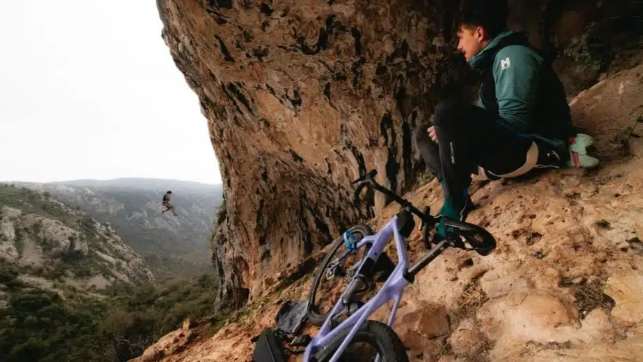Ein Mann mit einem lila Mountainbike sitzt in einer felsigen Umgebung auf dem Boden und blickt in die Ferne, wo ein anderer Mensch an einem Seil von einer Felswand abseilt.