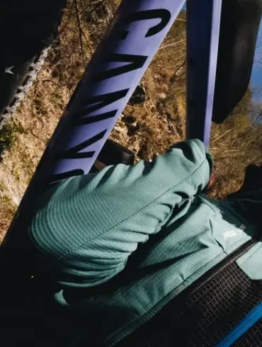 Nahaufnahme einer Frau mit Helm, die auf einem Fahrrad sitzt und ein grün-blaues Longsleeve von Millet trägt, im Hintergrund ein Fahrradrahmen mit der Aufschrift "CANYON".