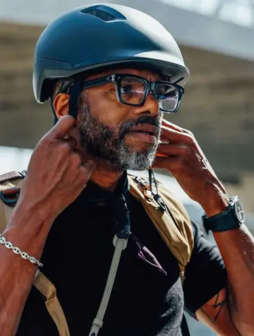 Ein Mann mit Bart und Brille setzt einen schwarzen Fahrradhelm auf und verstellt die Riemen. Der Helm ist für den urbanen Gebrauch konzipiert.