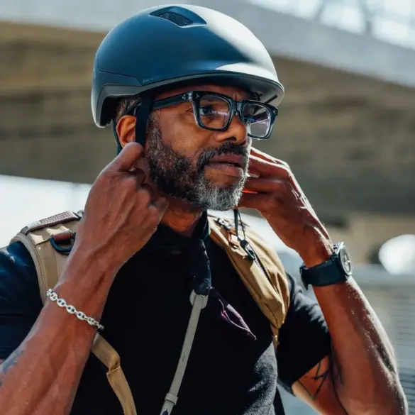 Ein Mann mit Bart und Brille setzt einen schwarzen Fahrradhelm auf und verstellt die Riemen. Der Helm ist für den urbanen Gebrauch konzipiert.