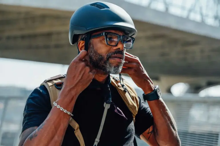 Ein Mann mit Bart und Brille setzt einen schwarzen Fahrradhelm auf und verstellt die Riemen. Der Helm ist für den urbanen Gebrauch konzipiert.