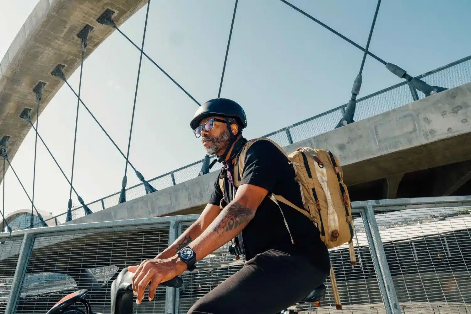 Mann mit schwarzem Fahrradhelm und Brille fährt Fahrrad unter einer Betonbrücke mit Stahlseilen, er trägt einen beigefarbenen Rucksack.