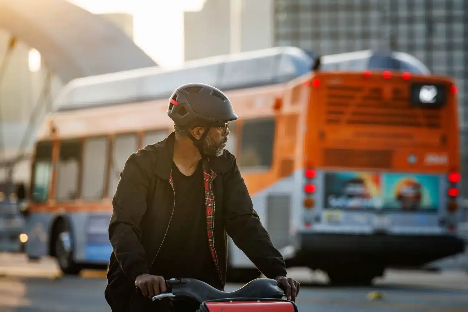 Ein Mann mit einem dunklen City-Helm, Brille und einem karierten Hemd unter einer Jacke blickt nach rechts, während er auf einem Fahrrad sitzt. Im Hintergrund ist ein orange-grauer Bus zu sehen, der in einer städtischen Umgebung fährt.