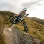 Ein Mountainbiker in Aktion auf seinem Pivot Firebird Bike, das über einen schmalen, erdigen Trail in einer grasbewachsenen Landschaft fährt. Der Fahrer befindet sich in der Luft, als ob er gerade einen Sprung gemeistert hätte. Die Sonne scheint im Hintergrund und erzeugt ein warmes Licht.