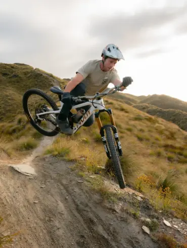 Ein Mountainbiker in Aktion auf seinem Pivot Firebird Bike, das über einen schmalen, erdigen Trail in einer grasbewachsenen Landschaft fährt. Der Fahrer befindet sich in der Luft, als ob er gerade einen Sprung gemeistert hätte. Die Sonne scheint im Hintergrund und erzeugt ein warmes Licht.