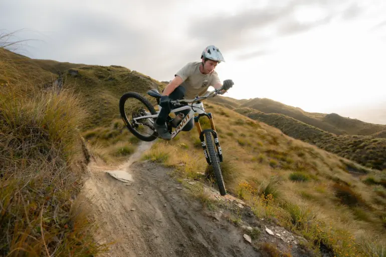 Ein Mountainbiker in Aktion auf seinem Pivot Firebird Bike, das über einen schmalen, erdigen Trail in einer grasbewachsenen Landschaft fährt. Der Fahrer befindet sich in der Luft, als ob er gerade einen Sprung gemeistert hätte. Die Sonne scheint im Hintergrund und erzeugt ein warmes Licht.