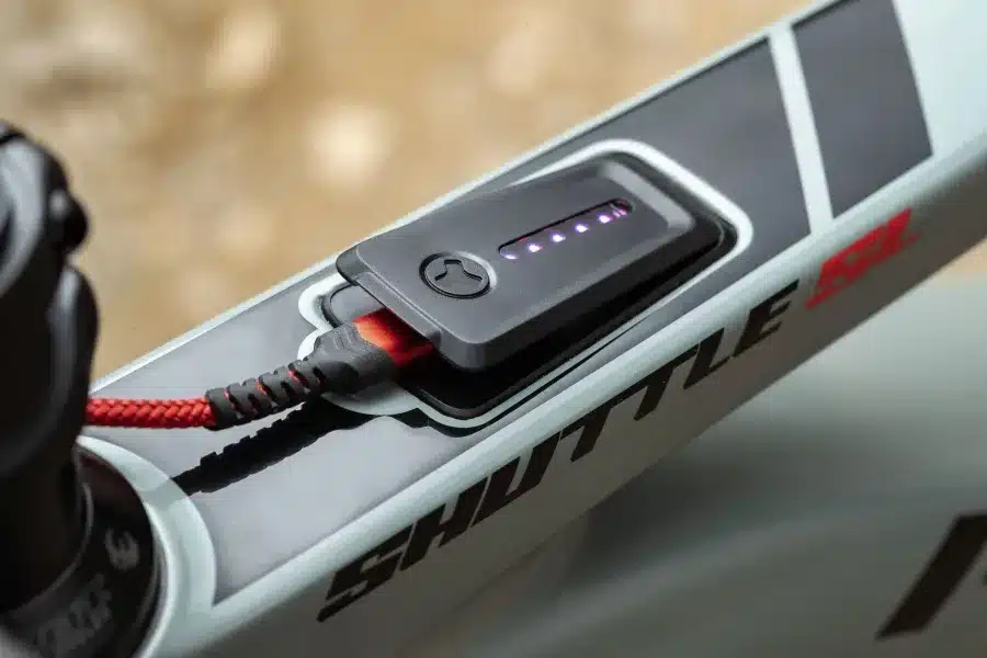 Nahaufnahme eines modernen Fahrradrahmens mit einem daran befestigten Akku, der über ein geflochtenes rotes USB-Kabel geladen wird. Auf dem Akku sind mehrere Ladeanzeigen-LEDs sichtbar, die leuchten.