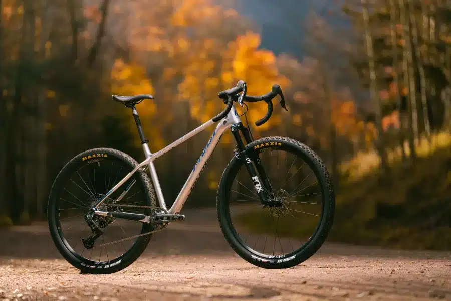 Ein silbernes Pivot LES SL Drop Bar Hardtail-Fahrrad mit Rennlenker steht auf einem Schotterweg, umgeben von Bäumen mit herbstlichem Laub.