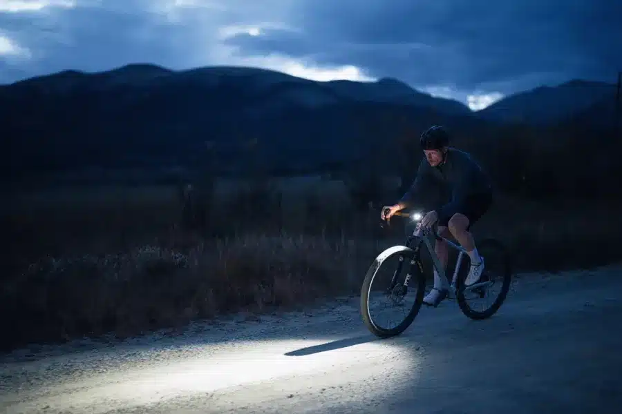 Ein Mann fährt mit einem Dropbar-Hardtail-Fahrrad nachts auf einem Schotterweg, beleuchtet von einem starken Scheinwerfer.
