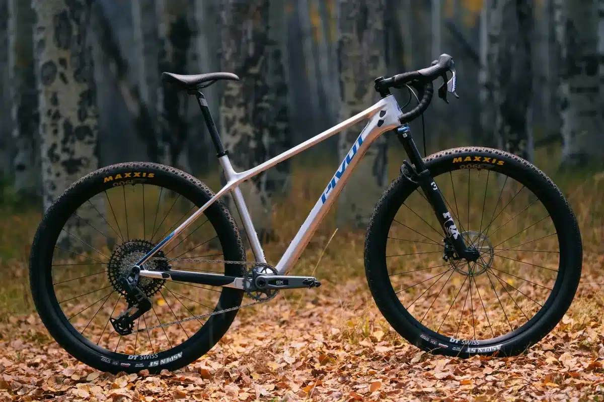 Ein weißes Pivot LES SL Drop Bar Fahrrad mit blauen Akzenten steht auf einem Waldboden bedeckt mit Herbstlaub.