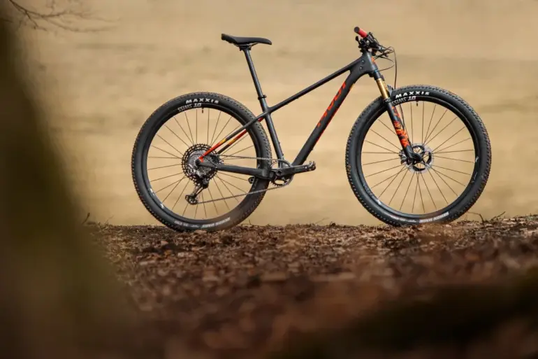 Ein schwarzes Pivot Les SL Cross-Country-Hardtail steht auf erdigen Blättern, das Vorderrad zeigt eine goldene Federgabel.