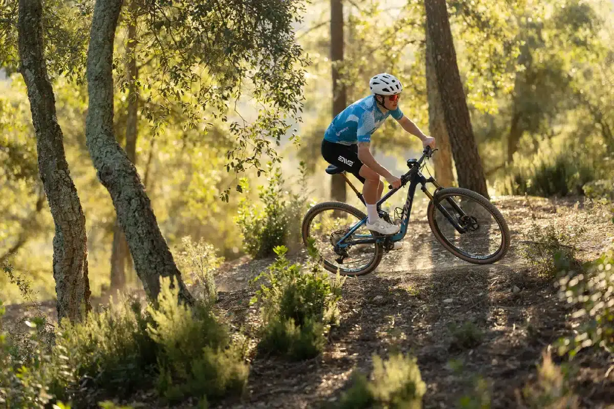 Ein Mountainbiker fährt auf einem schmalen Waldweg bergab. Die Sonne scheint durch die Bäume und wirft Schatten auf den Weg.