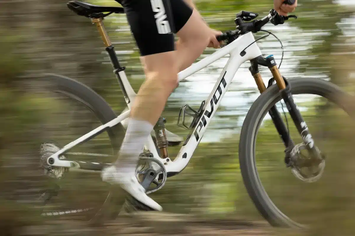 Ein weißes Pivot Mach 4 SL Mountainbike wird von einer Person gefahren, die auf einem bewaldeten Trail unterwegs ist. Der Fokus liegt auf der Bewegung und Dynamik des Rades, während der Hintergrund verschwommen ist.