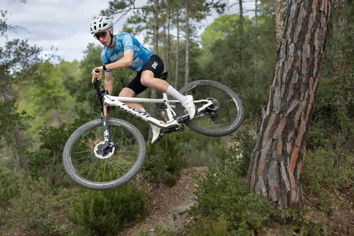 Ein Mountainbiker in voller Montur springt mit einem weißen Pivot Mach 4 SL Mountainbike über einen Waldweg.